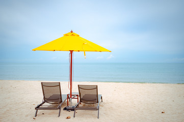 Beach chairs under a yellow umbrella on beach with soft blue sky