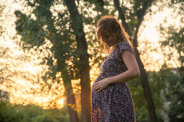 A happy pregnant woman standing in the city park holding her arms around the stomach.