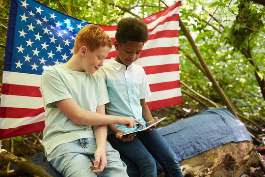 Portrait of two teenage boys using digital tablet outdoors while hiding under branches of big tree in forest or playing in back yard