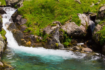 small waterfall, light blue water surrounder by grass and several rocks