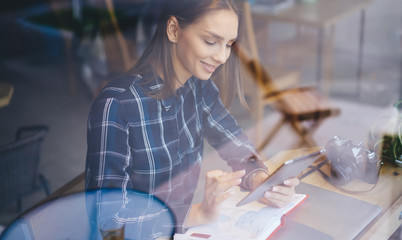 Young smiling hipster girl chatting with followers and sharing news in social networks on digital tablet connected to 4G internet.Attractive female watching funny video on gadget sitting indoors