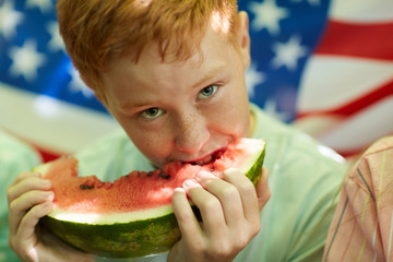 Close up portrait of freckled red-haired boy holding watermelon and looking at camera with American flag in background, copy space
