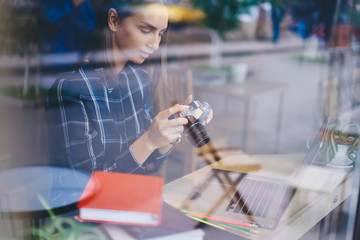 Pensive female journalist making settings on vintage camera preparing for taking pictures while working on article using laptop computer, hipster girl spending time on hobby testing old camera