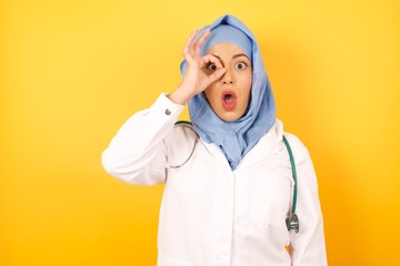 Young arab doctor woman wearing medical uniform standing over yellow background  doing ok gesture shocked with surprised face, eye looking through fingers. Unbelieving expression.