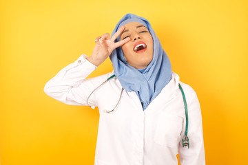 Young arab doctor woman wearing medical uniform standing over yellow background  Doing peace symbol with fingers over face, smiling cheerful showing victory