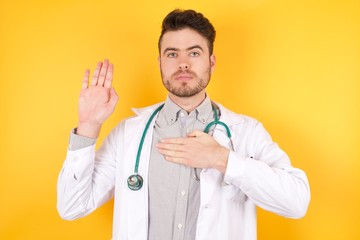 Young Caucasian doctor man wearing medical uniform, standing over isolated yellow background Swearing with hand on chest and open palm, making a loyalty promise oath