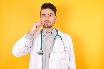 Young Caucasian doctor man wearing medical uniform, standing over isolated yellow background mouth...