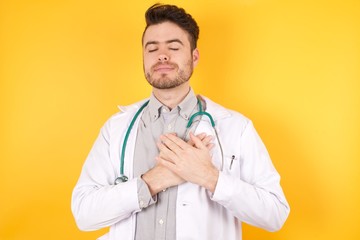 Young Caucasian doctor man wearing medical uniform over isolated yellow background smiling with hands on chest with closed eyes and grateful gesture on face. Health concept.