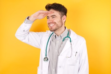 Young Caucasian doctor man wearing medical uniform, over isolated yellow background very happy and smiling looking far away with hand over head. Searching concept.