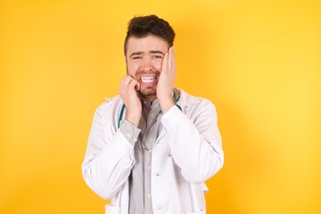 Doleful desperate crying doctor man wearing medical uniform, being in depression after quarrel with someone, looks stressfully, frowns face, isolated over yellow background. 