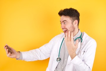 Wow!! excited handsome doctor man wearing medical uniform, showing mobile phone with open hand gesture isolated on yellow background