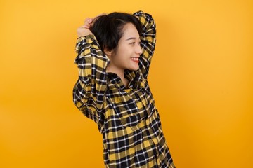 Young asian woman with short hair wearing plaid shirt standing over yellow background stretching arms, relaxed position.