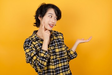 I wanna buy it! Photo of funny Young asian woman with short hair wearing plaid shirt standing over yellow background holding open palm new product 