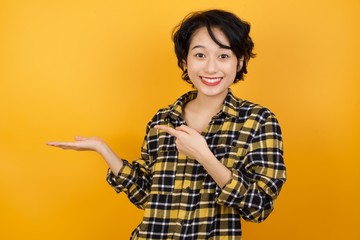 Close up photo of cute Young asian woman with short hair wearing plaid shirt standing over yellow background holding hand showing advertisement