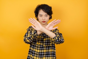Young asian woman with short hair wearing plaid shirt standing over yellow background has rejection expression crossing arms and palms doing negative sign, angry face.