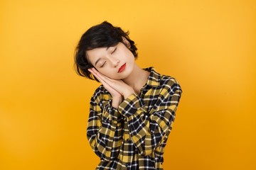 Young asian woman with short hair wearing plaid shirt standing over yellow background sleeping tired dreaming and posing with hands together while smiling with closed eyes.