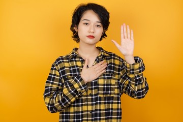 Young asian woman with short hair wearing plaid shirt standing over yellow background Swearing with hand on chest and open palm, making a loyalty promise oath
