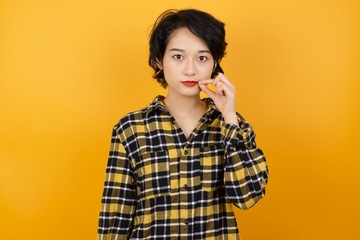 Young asian woman with short hair wearing plaid shirt standing over yellow background mouth and...