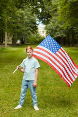 Vertical full length portrait of smiling red haired boy carrying American flag while standing outdoors in park, copy space