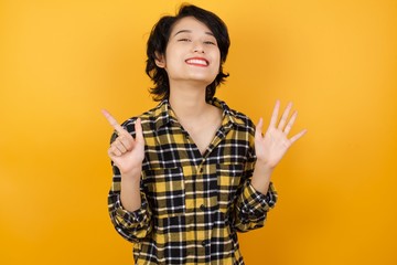 Young asian woman with short hair wearing plaid shirt standing over yellow background showing and pointing up with fingers number seven while smiling confident and happy.