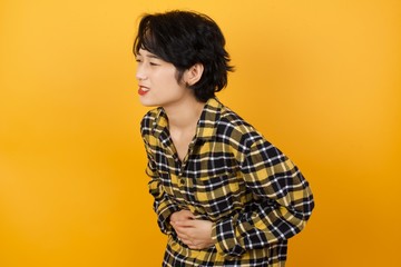 Young beautiful Asian woman wearing plaid shirt over yellow background with hand on stomach because nausea, painful disease feeling unwell. Ache concept.