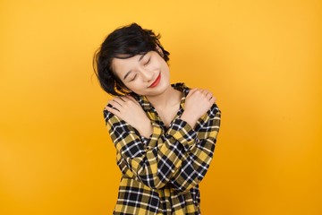 Young beautiful Asian woman wearing plaid shirt over yellow background Hugging oneself happy and...