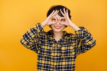 Young beautiful Asian woman wearing plaid shirt over yellow background doing ok gesture like binoculars sticking tongue out, eyes looking through fingers. Crazy expression.