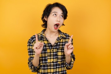 Young beautiful Asian woman wearing plaid shirt over yellow background amazed and surprised looking up and pointing with fingers and raised arms.