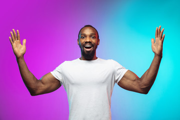 Naklejka premium Showing a huge. African-american young man's portrait on gradient studio background in neon. Beautiful male model in casual style, white shirt. Concept of human emotions, facial expression, sales, ad.