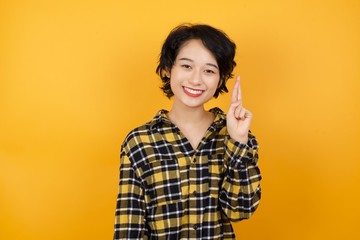Young asian woman with short hair wearing plaid shirt standing over yellow background pointing up with fingers number ten in Chinese sign language Shi