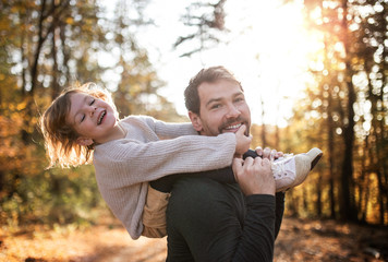 Mature father giving piggyback ride to small daughter on a walk in autumn forest.