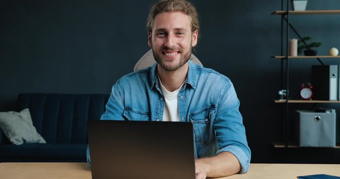 Young Successful Freelancer is sitting at Home Office, smiling at the Camera. Portrait of Satisfied Young Man is working on his Laptop at Home, looking directly in Shoot. Casual Style. Freelance.