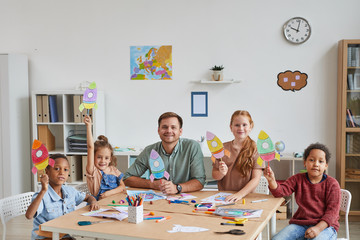 Portrait of smiling male teacher with multi-ethnic group of kids showing pictures of space rockets...