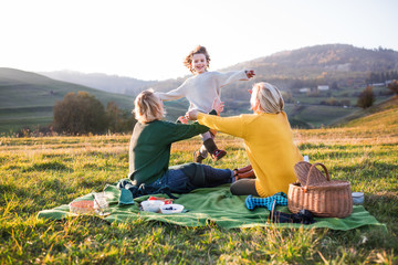 Small girl with mother and grandmother having picnic in nature at sunset.