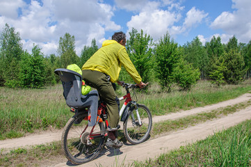 Dad in bright clothes rides his little daughter in a bicycle seat on a bicycle along a dirt road through the woods