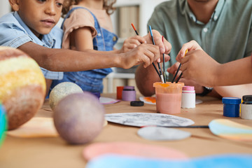 Close up of multi-ethnic group of kids holding brushes and painting planet model while enjoying art and craft lesson in school or development center, copy space