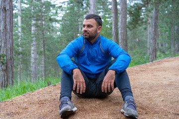 Naklejka na ściany i meble Young athlete in blue sports wear stretching in woods. Taking rest after jogging.