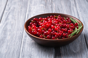 Fresh sweet red currant berries with rosemary leaves in wooden bowl, grey wood background, angle view