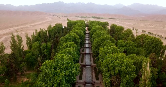 Aerial rising drone shot moves up the fresh garden with green trees, stream and beautiful palace building with a desert and mountain landscape in Iran