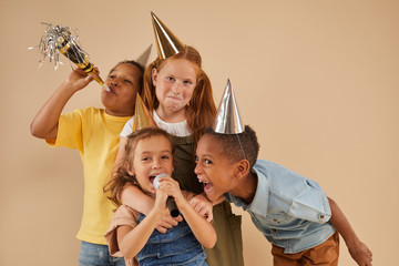 Waist up portrait of diverse group of children wearing party hats screaming to microphone while...