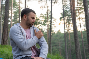 Pretty young beard man having pain in his shoulder. Sitting in forest,surrounded with trees and beautiful nature. 