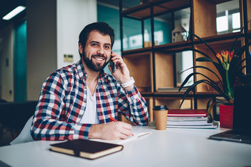 Positive bearded man with mobile phone calling to friends using application, smiling happy young hipster guy looking away while talking via modern cellular indoors, technology and communication