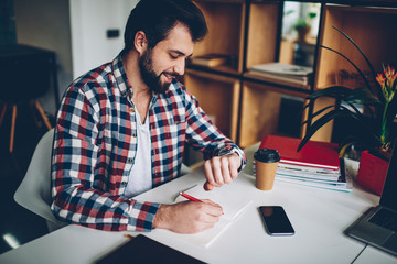 Caucasian bearded young man looking at wristwatch while working indoors, hipster guy 20s learning and checking time on wearable clock while writing creative ideas for article in notepad at desktop