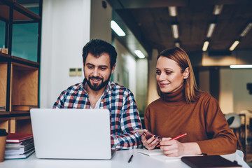 Two cheerful persons watching webinar via laptop online sitting at coworking space, smiling hipster...