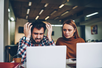 Angry male IT developer disappointed of work application at modern laptop sitting with female business colleague at coworking space, hipster guy feeling stress from watching video on netbook