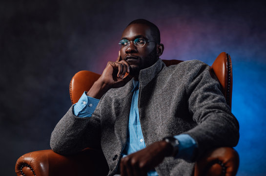 A blackman dressed in expensive and fashionable quality clothes. Leather nice armchair with african guy and multicolored background behind.