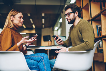 Young hipsters students holding cellulars and communicated enjoying time at university campus, two friends using mobile phones and chatting online via application while discussing day indoors