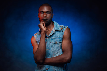 Fototapeta premium Fashionable attractive black guy in jean jacket. An african person with blue foggy background behind. Urban style.