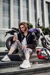 Obraz na płótnie Canvas Cool, beautiful and stylish girl in glasses in black frame and checkered trousers sits on the steps of the stairs near her purple motorcycle and looks at the screen of her mobile phone