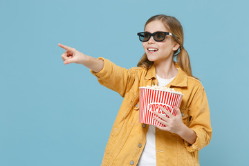 Fototapeta premium Pretty little kid girl 12-13 years old in 3d glasses isolated on blue background. Childhood lifestyle concept. People in cinema. Watching movie film hold bucket of popcorn pointing index finger aside.
